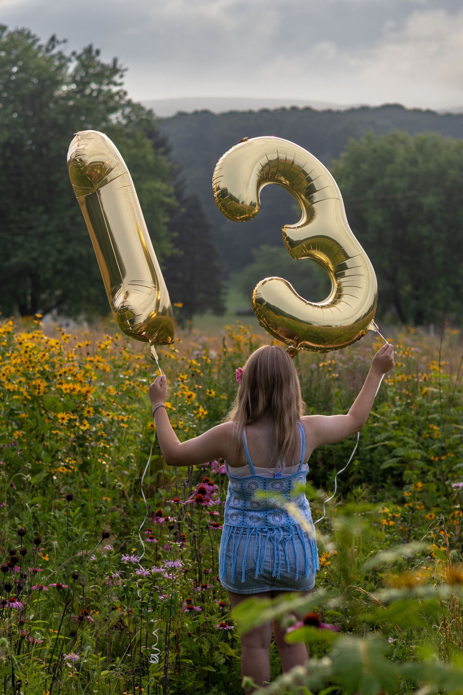 A girl stands in a wildflower meadow holding gold number 13 balloons above her head, celebrating her 13th birthday.