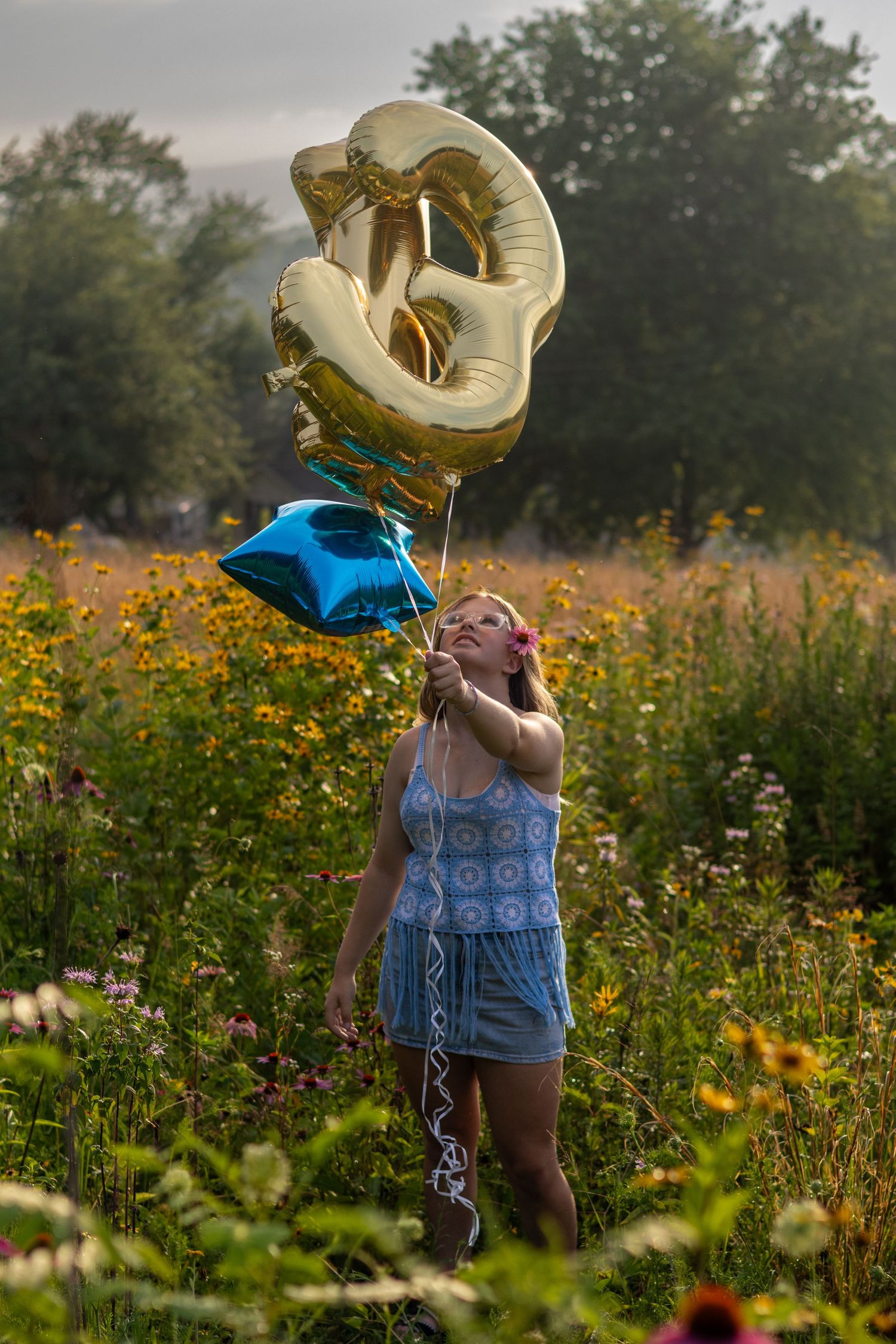 Young woman holding gold number and blue star balloons in a vibrant wildflower field on a misty day.