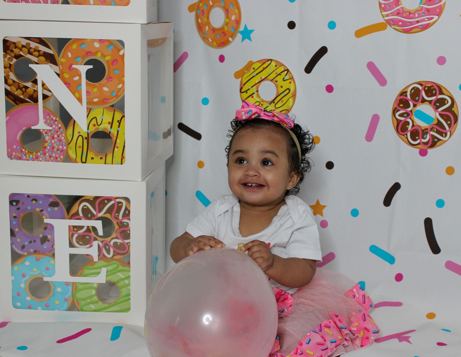 Baby girl smiles at donut-themed first birthday party with balloon, decorative blocks spelling ONE, and colorful donut backdrop.