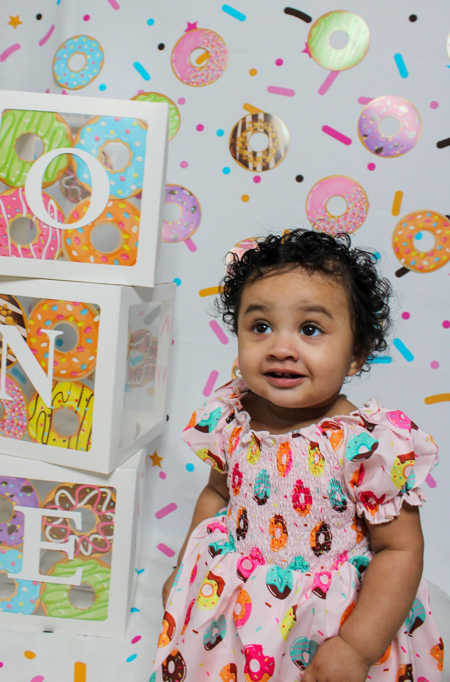 Cute baby girl in donut-print dress poses beside stacked ONE blocks at a colorful donut-themed first birthday party.