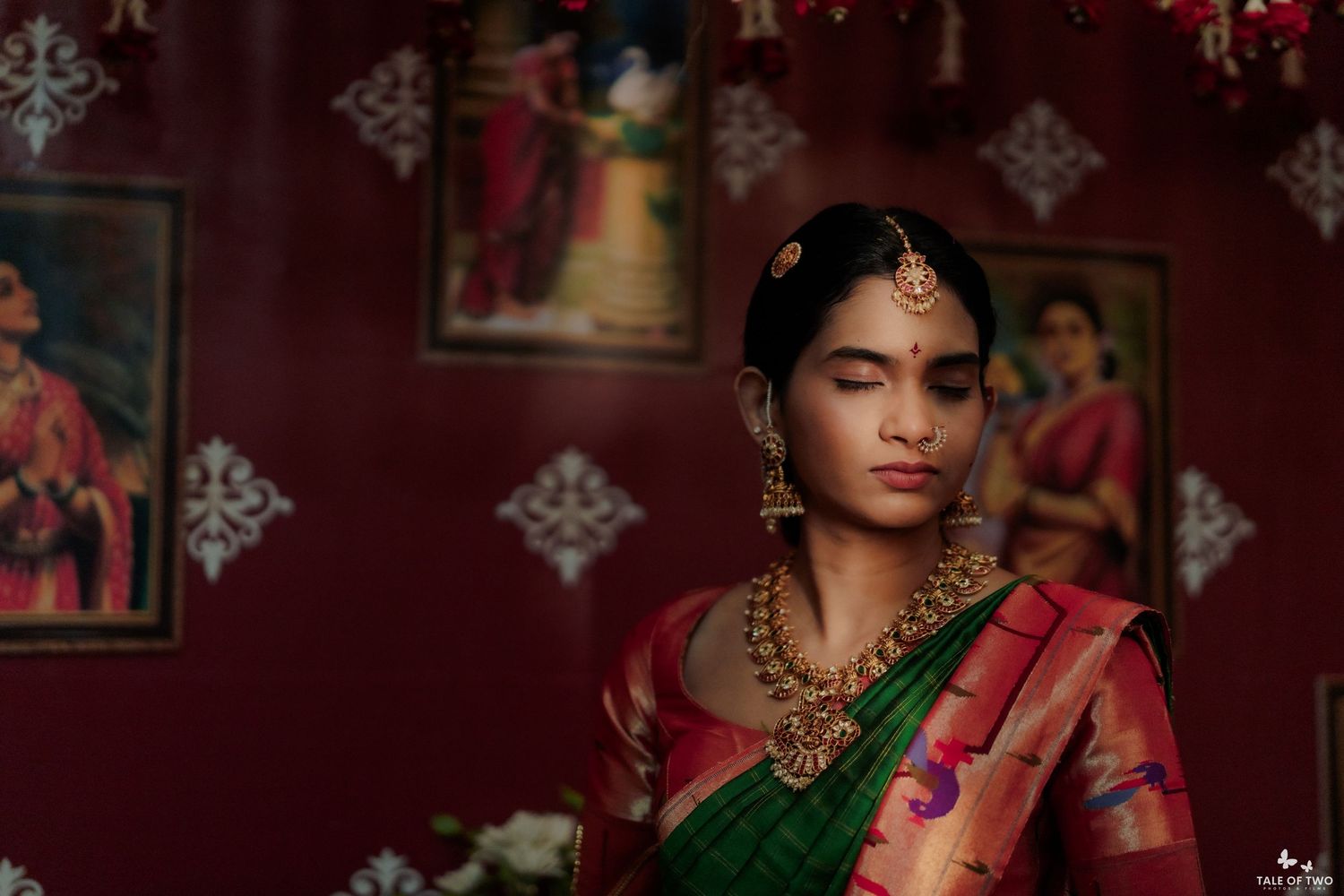 Indian bride in green pink Kanjivaram saree with gold temple necklace posing in decorated red room.