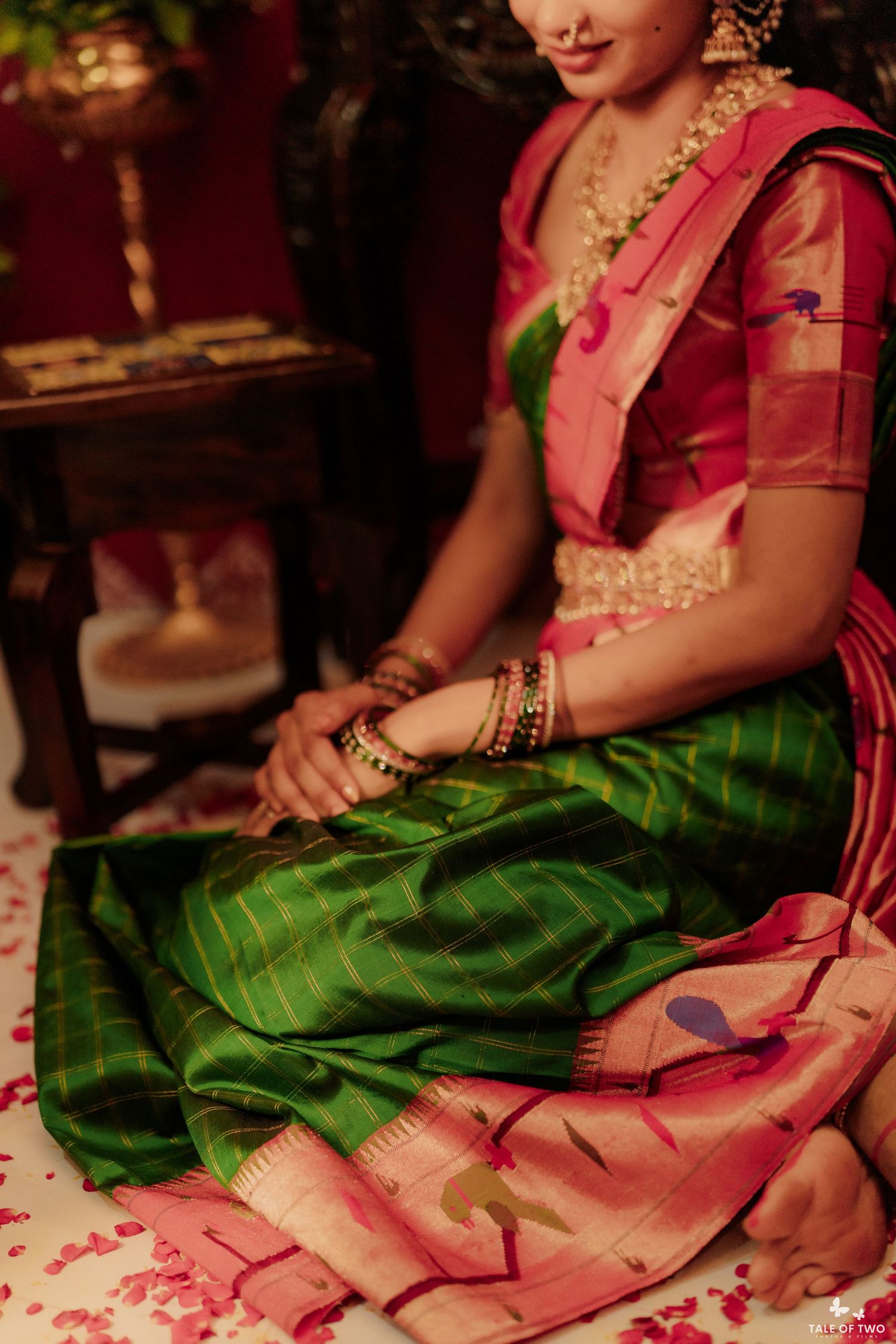 Woman in green and pink silk Kanjivaram saree with gold jewelry, seated gracefully on floor with rose petals.