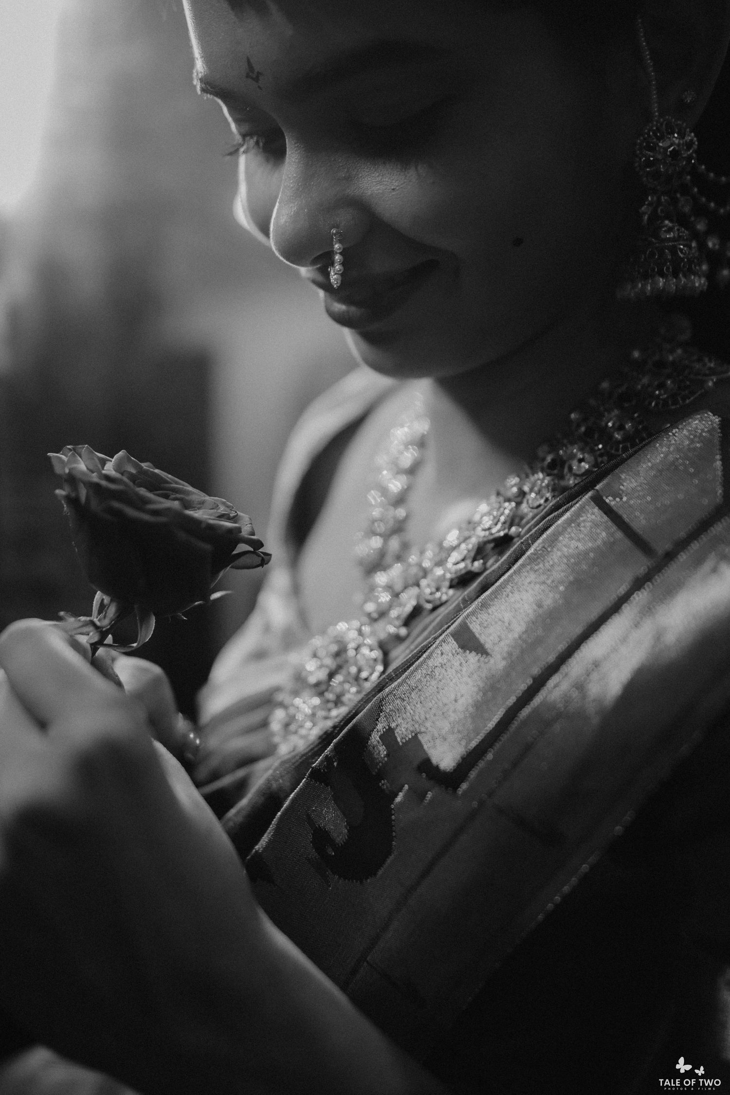 Black and white photo of a bride in traditional jewelry holding a rose and violin, smiling softly.