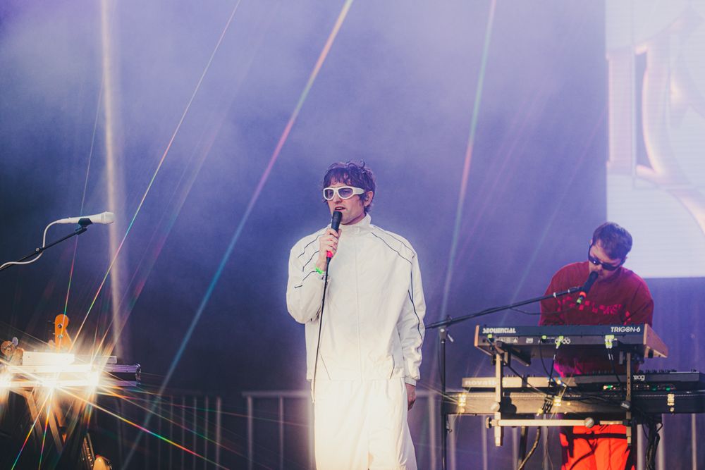 Singer in white suit performs on stage with laser lights while a musician plays keyboards in the background.