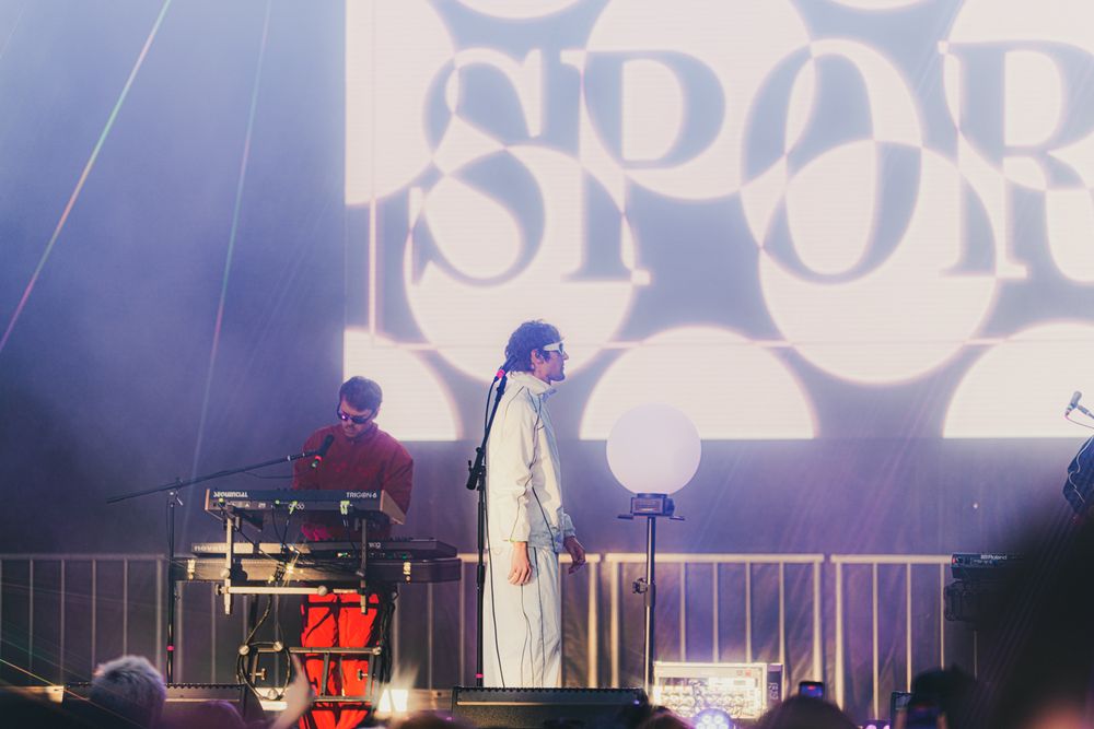 Two musicians perform on stage with a large projected backdrop displaying bold graphic text at an outdoor concert.