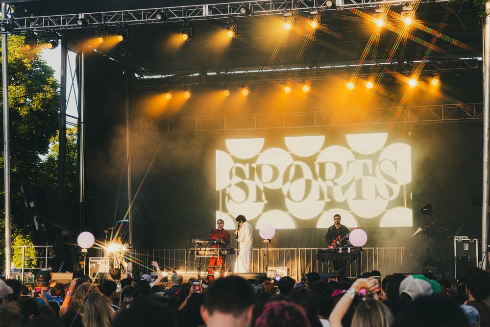 Band Sports performing live on a festival stage with yellow lights and a bold logo backdrop before a large crowd.
