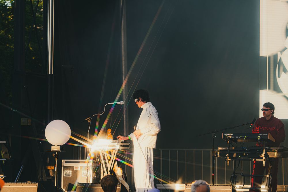 Performer in white outfit playing keyboard on outdoor stage with colorful lens flare at music festival.