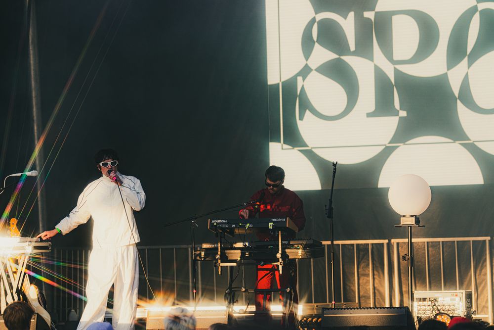 Two performers on stage at a music festival, one in white outfit rapping and another DJing with a large logo backdrop.