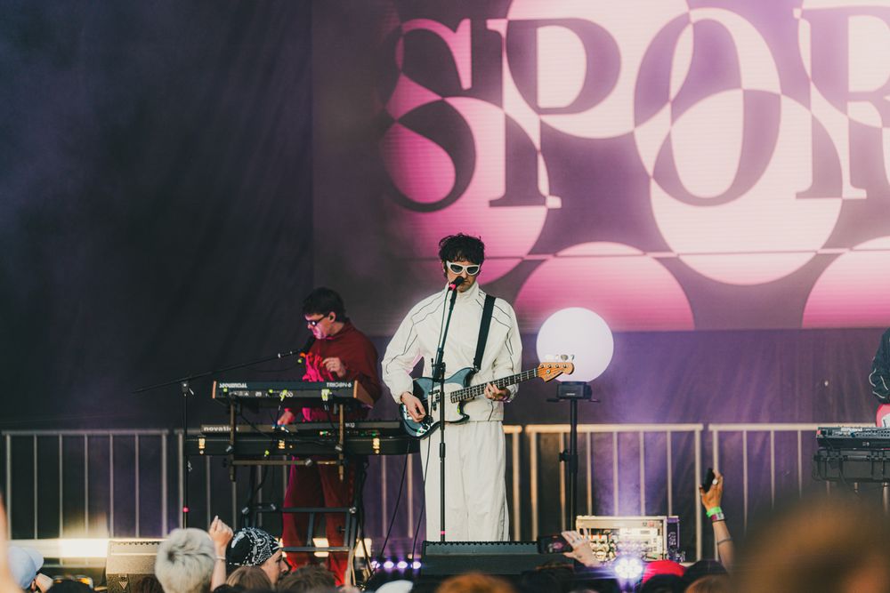 Band performing on outdoor festival stage, guitarist in white robe with sunglasses, DJ at keyboards, pink backdrop.