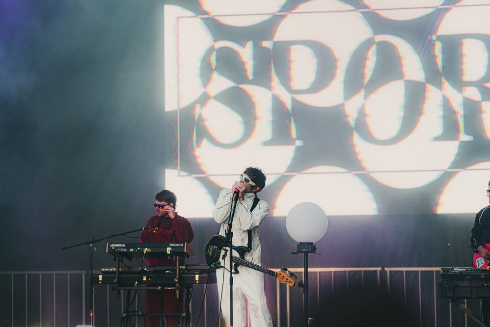 Singer performs on stage with keyboard player behind her, large SPOR logo glowing on screen in smoky concert venue.