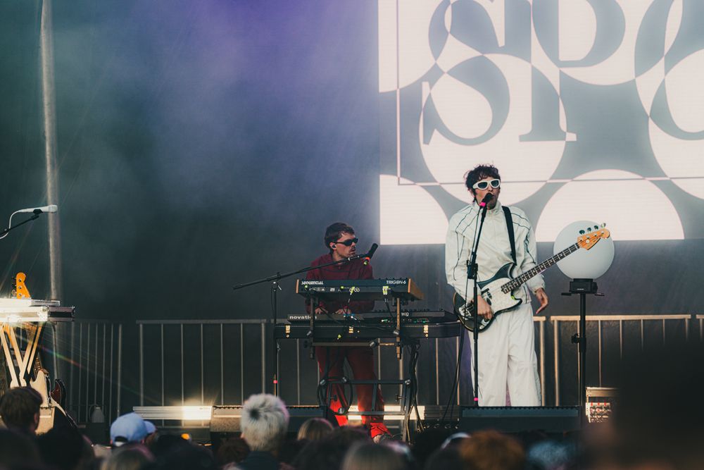 Two musicians perform on stage, one on keyboard and one with bass guitar, large circular SPOR logo lit behind them.