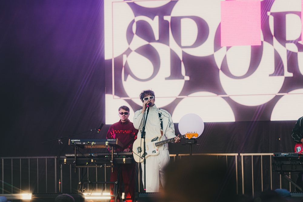 Musician in white suit playing guitar on stage with DJ behind keyboard and circular SP backdrop at live concert.