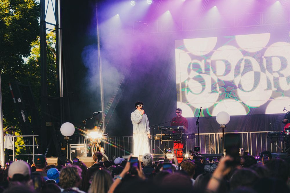 A performer in white on an outdoor festival stage with colorful lights and a large crowd at night.