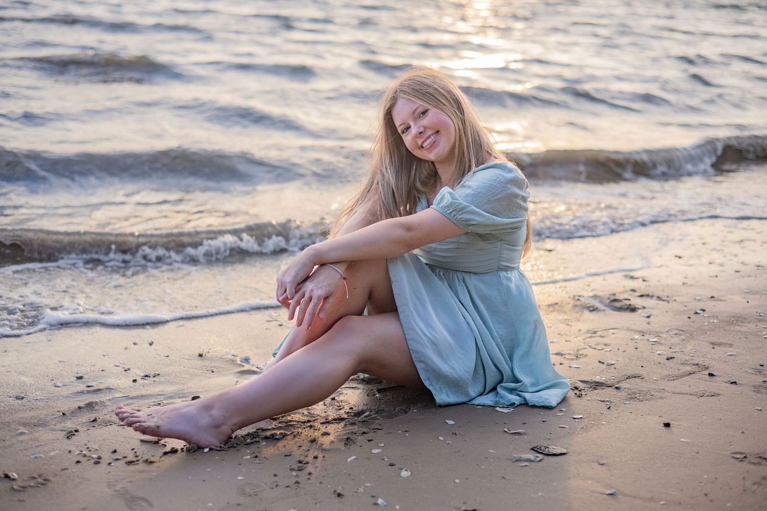 Person in a light blue dress sitting on a sandy beach at sunset with waves in the background.