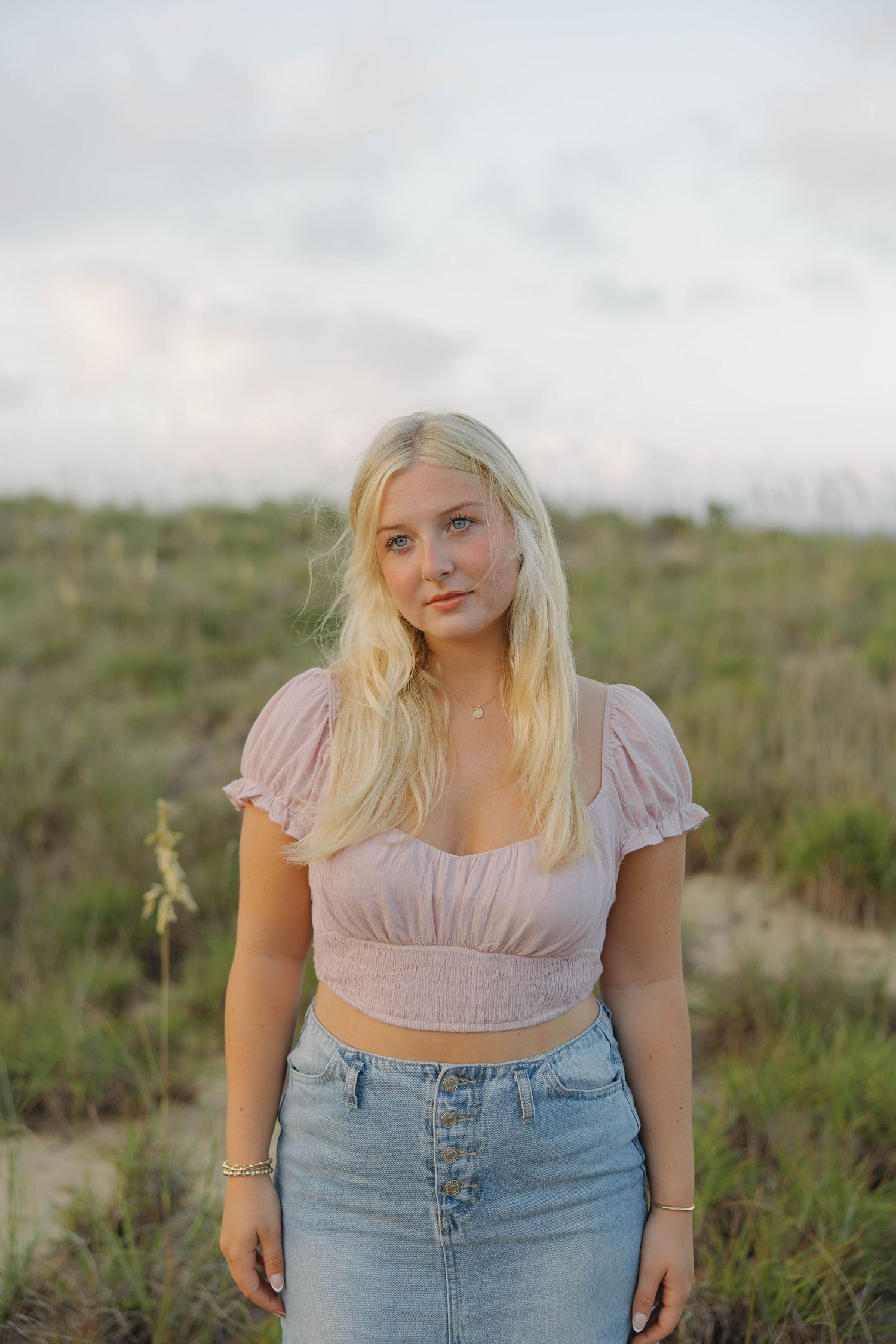 Young woman in crop top and high-waisted jeans poses in grassy field during sunset.