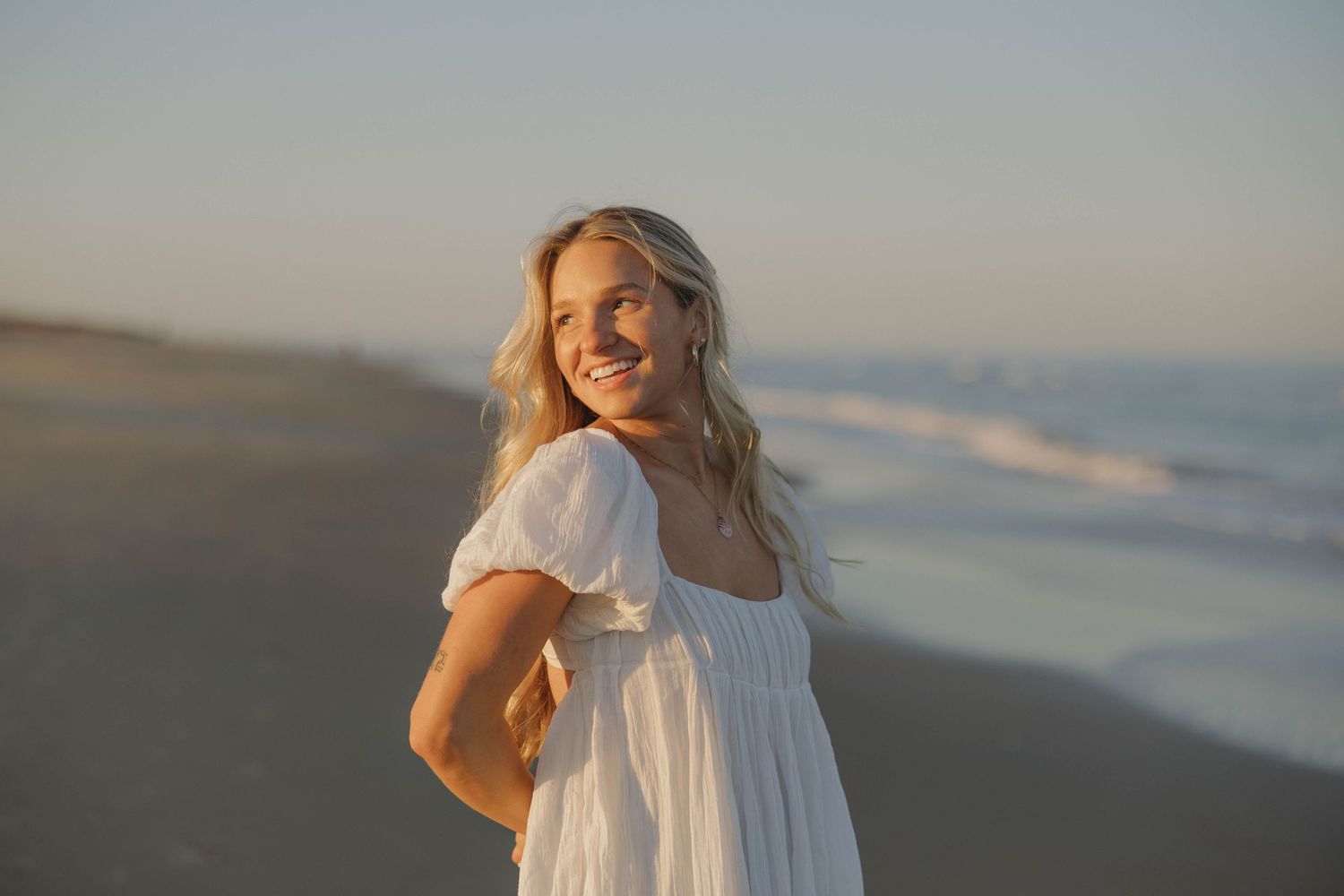 Woman in flowing white dress walking along beach at sunset with soft golden light and ocean waves.