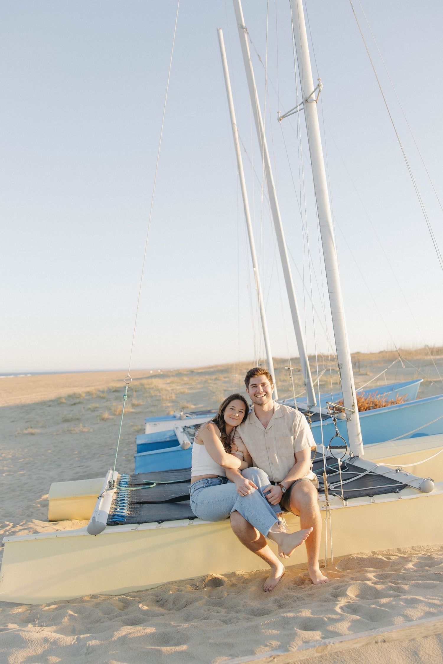 A couple sits together on a beached sailboat at sunset, with masts and rigging visible against the pale sky.