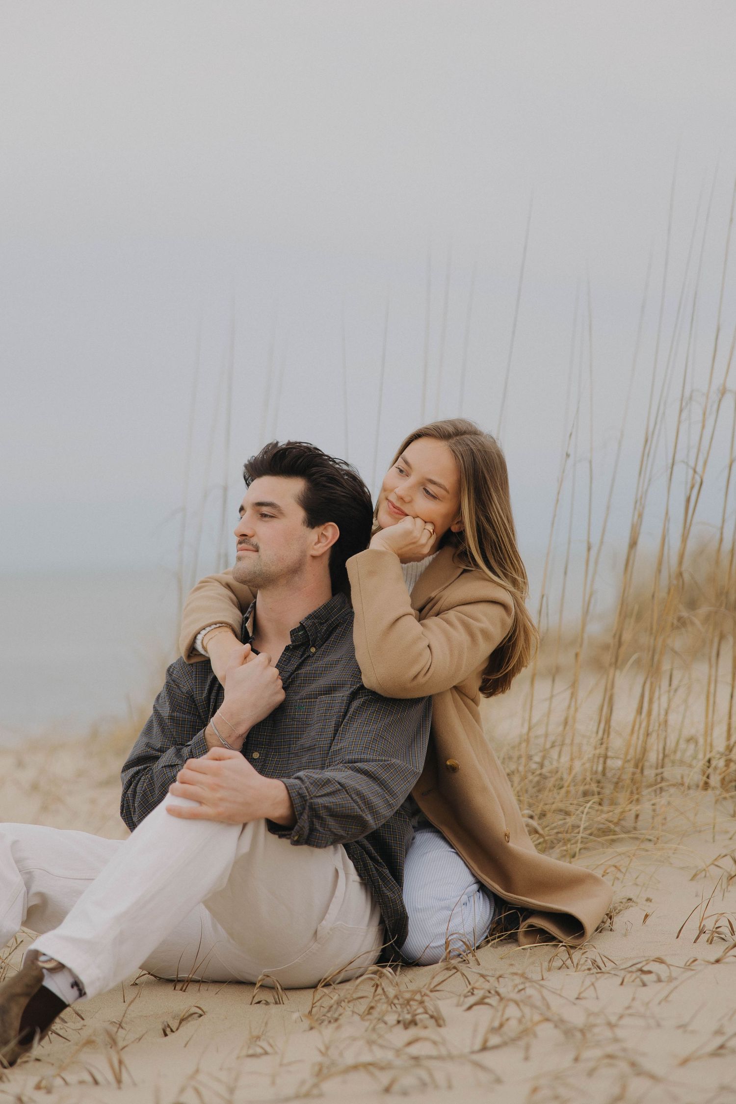 A couple shares tender moments while relaxing together on a sandy beach with tall grass in the background.