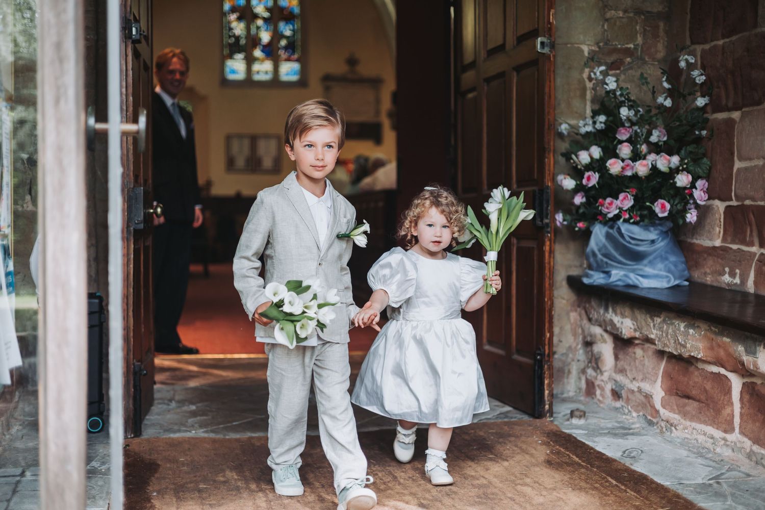 Two young children in formal attire hold flowers while standing in a church doorway during a wedding ceremony.