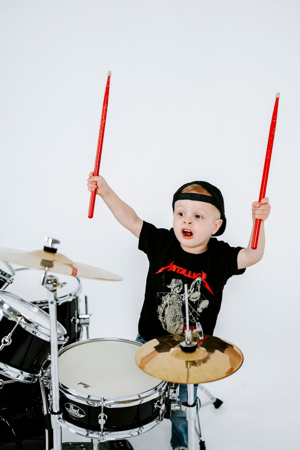 A young drummer in a black t-shirt plays energetically on a white drum kit while holding red drumsticks.
