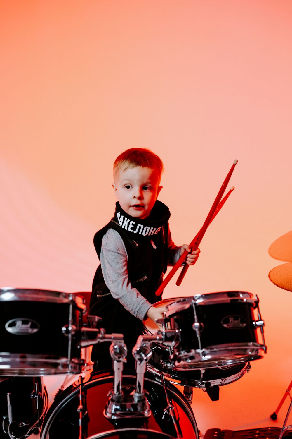 A young child plays enthusiastically on a drum set against a vibrant orange-tinted background.