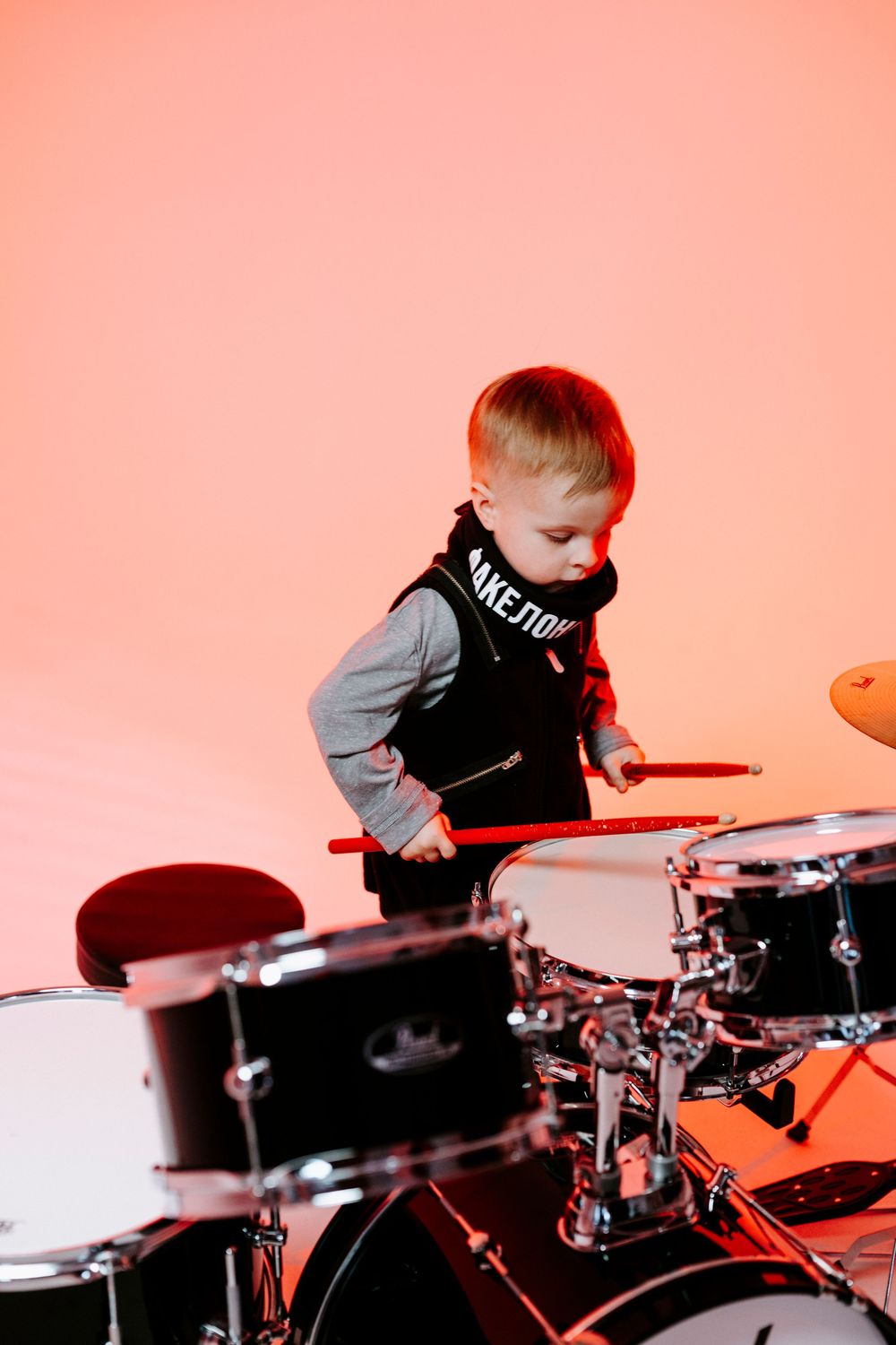 A young child in a black shirt plays an electronic drum set against a pink-tinted background.