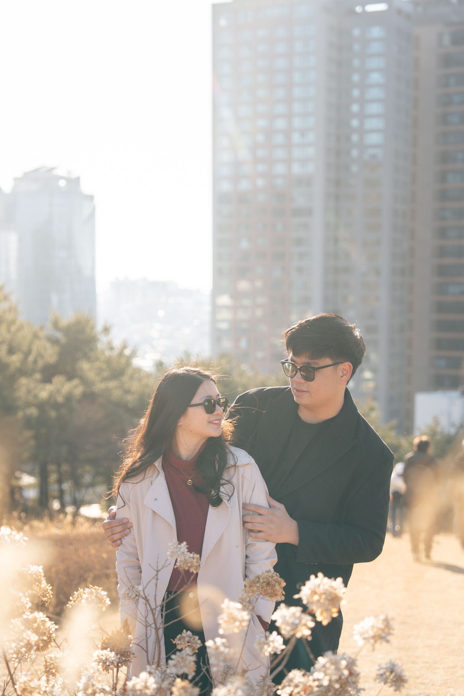 A couple in sunglasses embraces in a golden sunlit field with tall city buildings visible in the background.