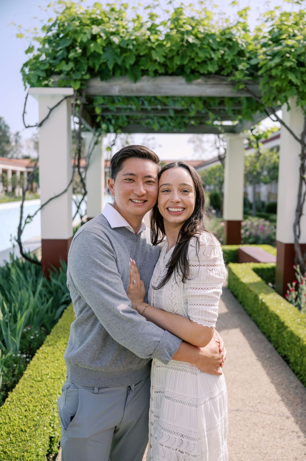 A couple embraces in a garden pathway under a vine-covered pergola with manicured hedges and landscaping.