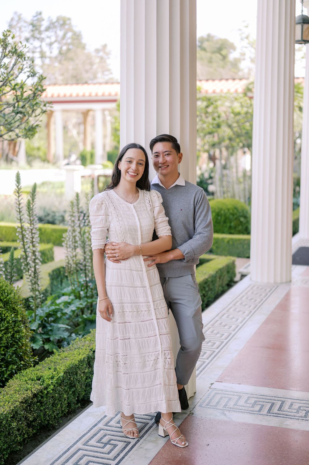 A couple poses together on an elegant columned porch with manicured gardens in the background.