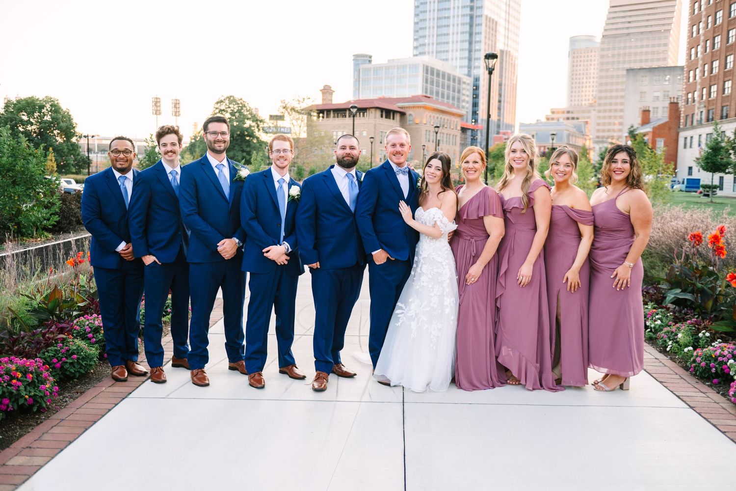 Wedding party in navy suits and mauve dresses poses together on a sunny day in an urban garden setting.