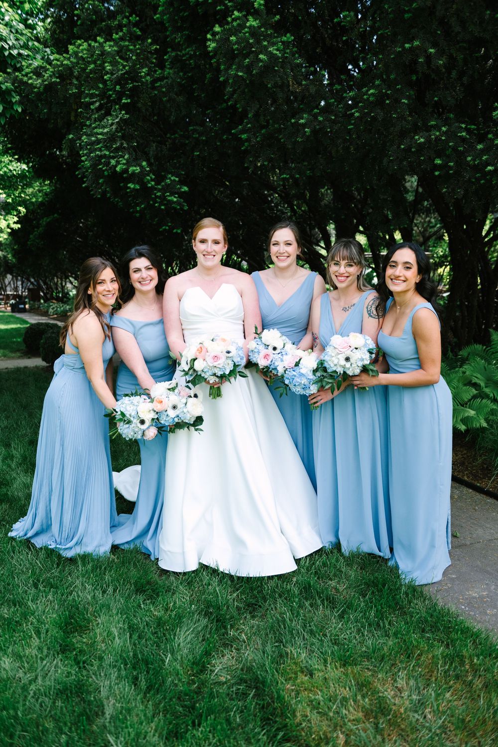 Bridal party in light blue bridesmaid dresses pose together on grassy lawn.