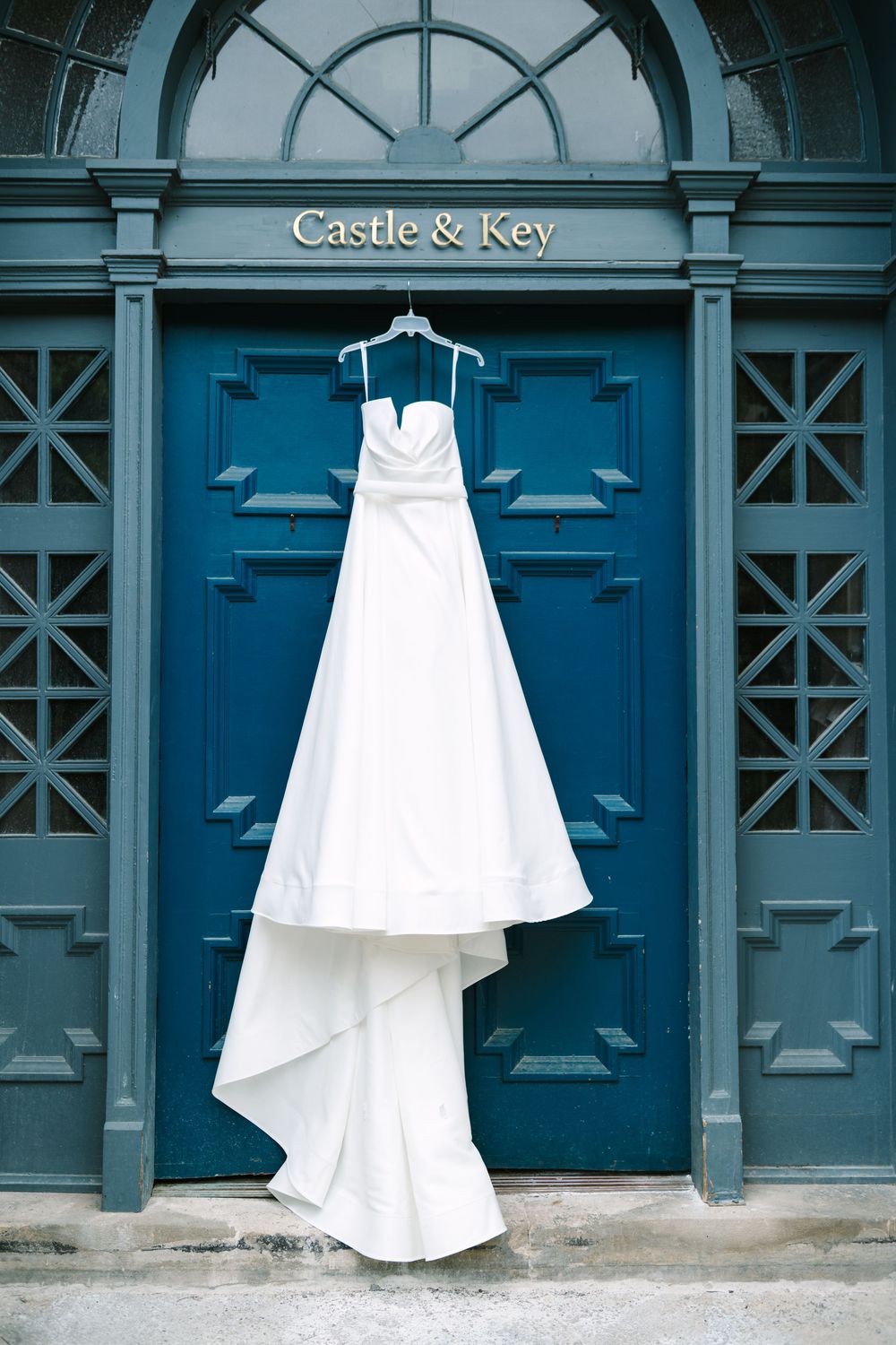 White wedding dress hanging against blue doors at Castle & Key distillery creates elegant contrast.