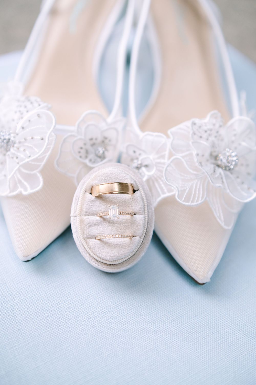 Wedding rings rest elegantly on white lace next to a pair of classic bridal shoes against a soft blue background.