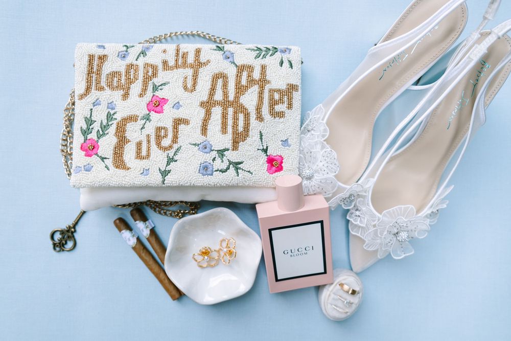 Wedding accessories including white heels, jewelry and gold lettered signage arranged on light blue surface.