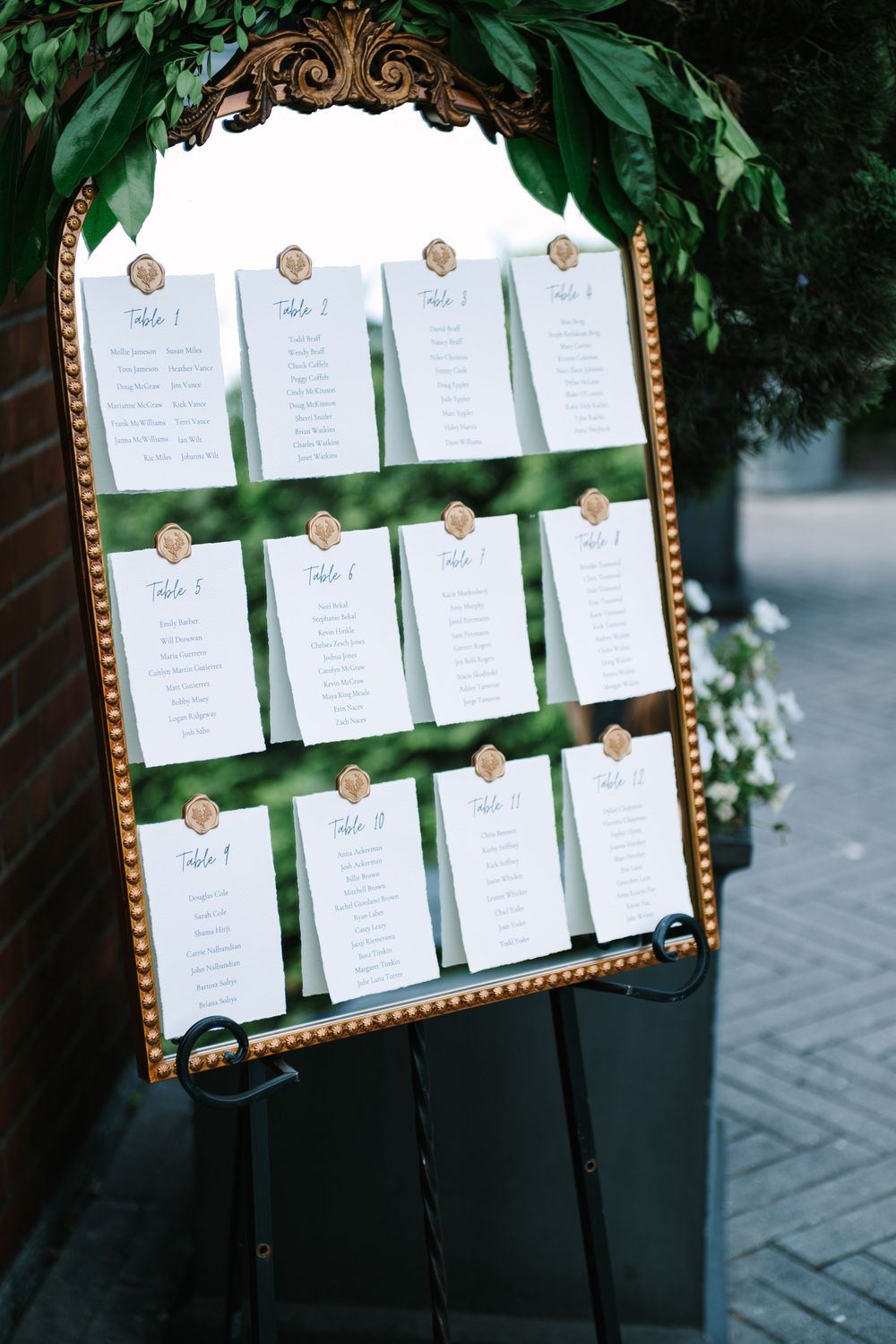 An ornate gold-framed seating chart with white cards arranged on green ribbons against a leafy backdrop.