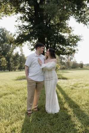 Couple in white outfits sharing a romantic moment under a large tree in a sunlit green park.