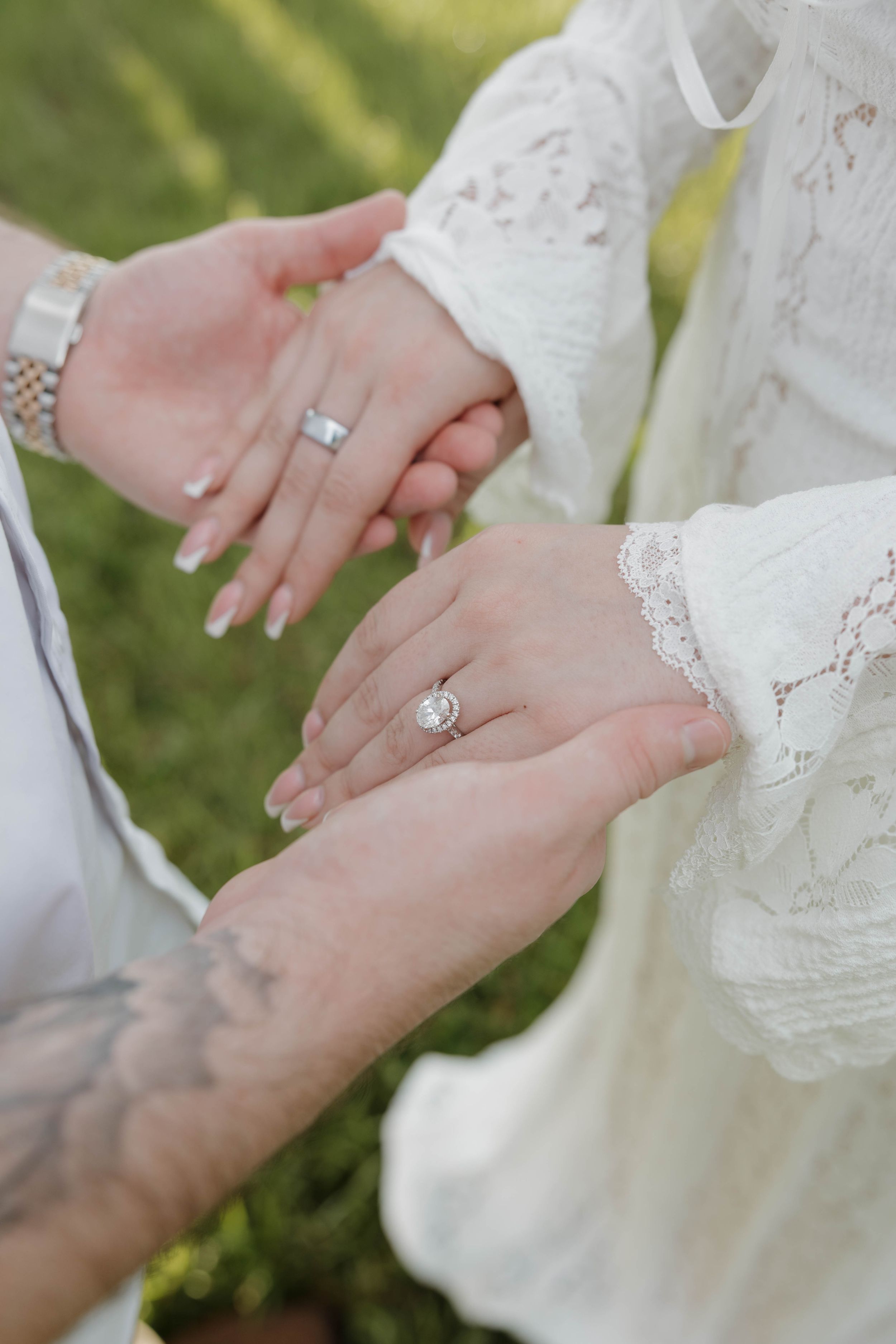 Bride and groom displaying wedding rings, bride wearing lace dress with diamond engagement ring, groom with tattoo and watch.