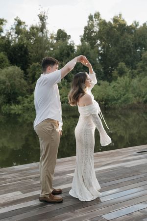 A couple dances on a wooden dock by a lake, the woman in a white lace gown and the man in a white shirt and khakis.