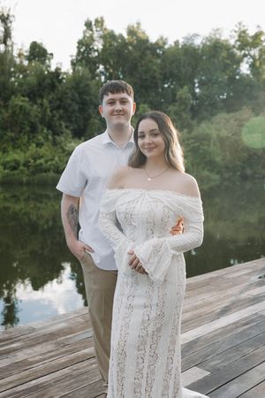 Couple posing on a wooden dock by a reflective lake, woman in white lace off-shoulder gown, man in white shirt.