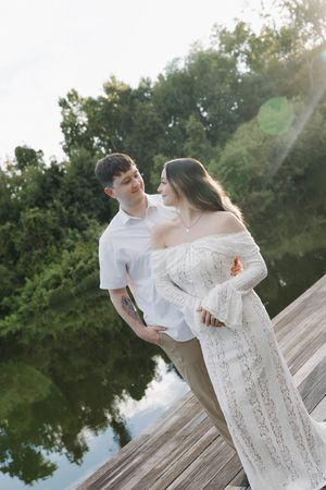 Couple in white outfits share a tender moment on a wooden dock by a serene lake surrounded by lush trees.