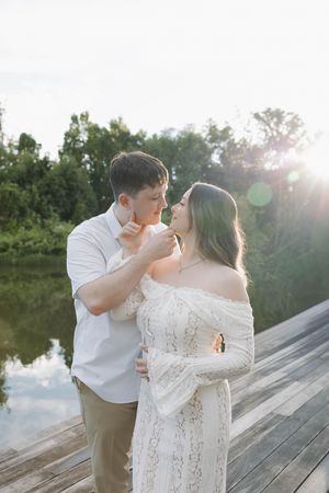Couple sharing a romantic moment on a wooden dock at sunset, woman in white lace dress, man in white shirt.