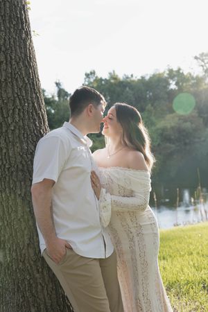 A couple shares a romantic moment leaning against a tree in a sunlit park, woman in white lace dress.