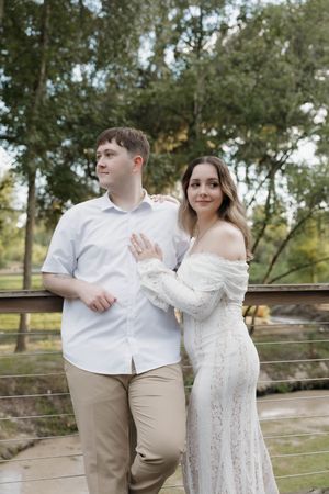 Couple posing on a bridge outdoors, woman in white lace dress embracing man in white shirt and khaki pants.