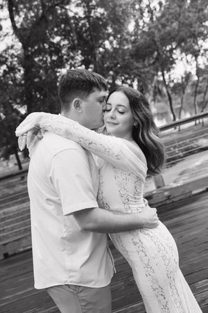 Black and white photo of a couple embracing outdoors, man kissing woman's cheek as she smiles in a lace dress.