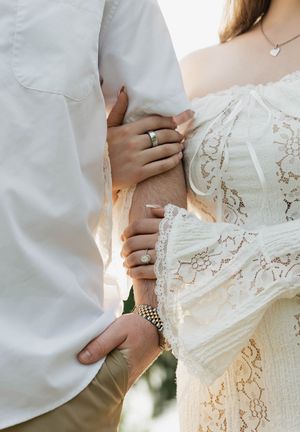 Close-up of a couple holding hands, showcasing engagement rings, lace dress, and bracelet in a romantic outdoor setting.