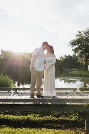 Couple shares a romantic forehead touch on a sunlit dock by a peaceful pond with golden hour backlight.