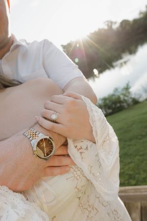 Close-up of a bride's diamond engagement ring and groom's gold watch as they embrace outdoors near a sunlit lake.