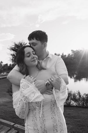 Black and white photo of a couple embracing near a lake at sunset, man kissing womans head while she smiles peacefully.