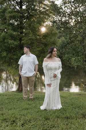 Couple in white attire standing by a serene pond surrounded by lush green trees at golden hour.