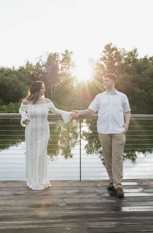 Couple holds hands on a wooden bridge at sunset, woman in white lace gown, man in white shirt and khakis.